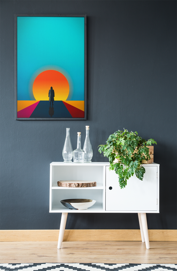 Framed artwork of a sunset with a person on a path above a white cabinet with decorative items.